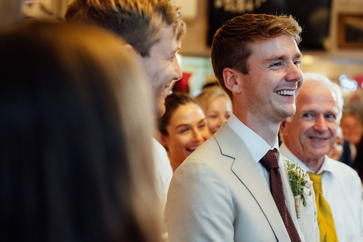 Smiling groom in a light gray suit and burgundy tie with a boutonniere stands among laughing wedding guests at a celebration.