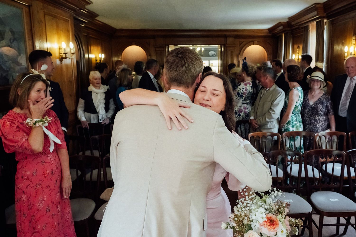 A groom in a light suit embraces a woman holding a bouquet during an emotional wedding ceremony as guests look on in a elegant paneled room.