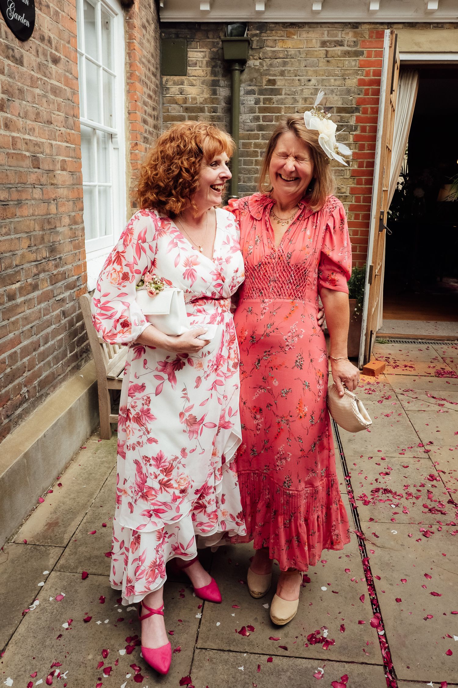 Two smiling women in floral summer dresses embrace outside a brick building, with pink petals scattered across the paving at a wedding.