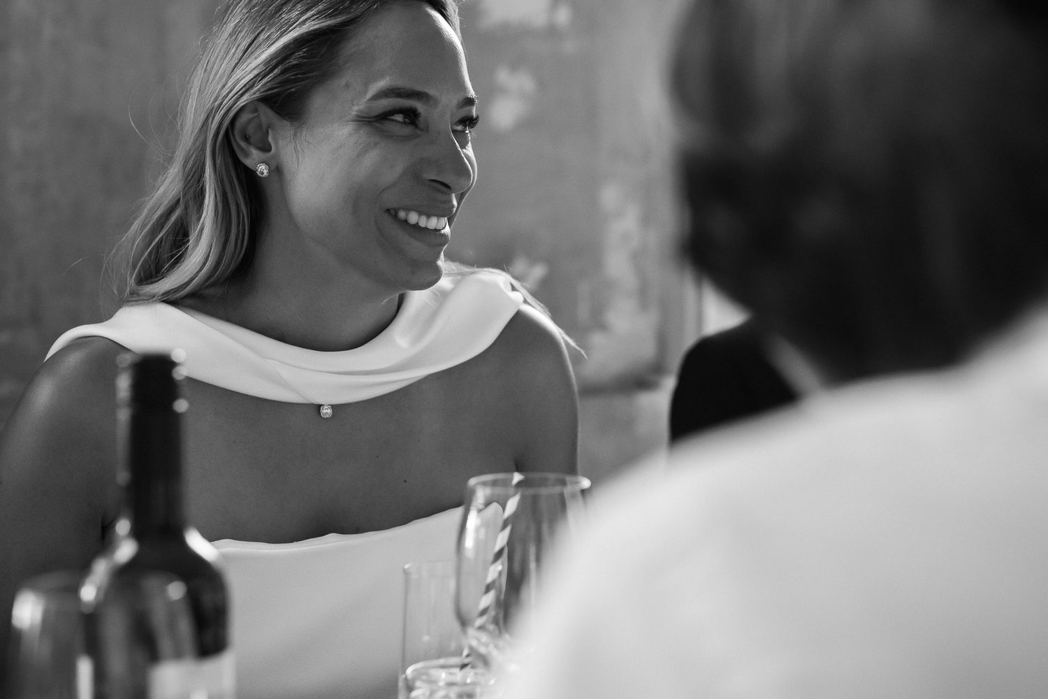 Smiling woman in a white off-the-shoulder dress laughs at a dinner table with a wine bottle and glasses in a black and white photo.