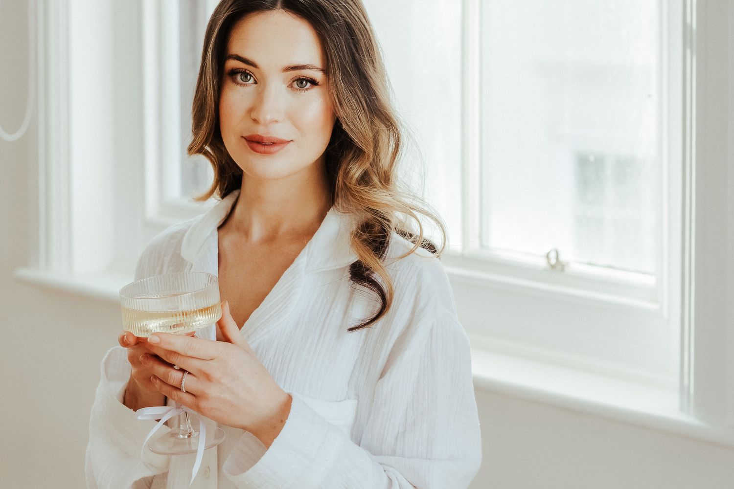 Woman in white robe sits by bright window holding coffee cup and smiling at morning light.