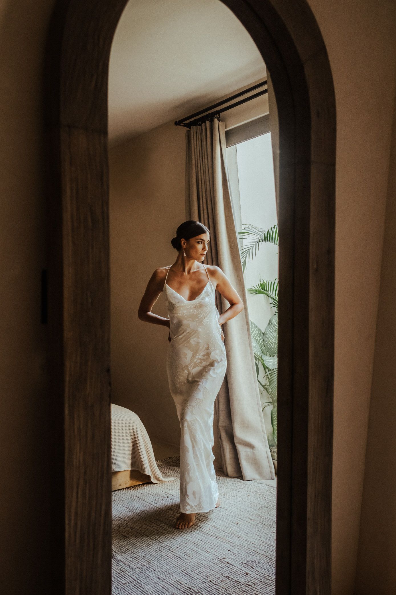 A bride in a white lace wedding dress poses elegantly near a window in a softly lit room.