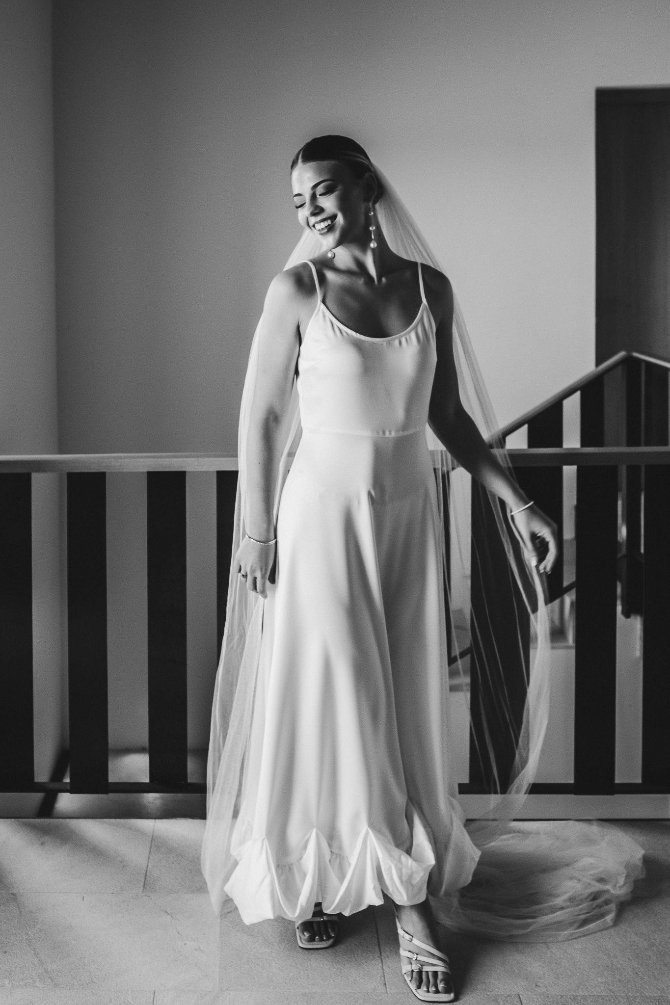 Black and white portrait of a bride in a simple elegant wedding dress and veil smiling while wearing casual sandals.