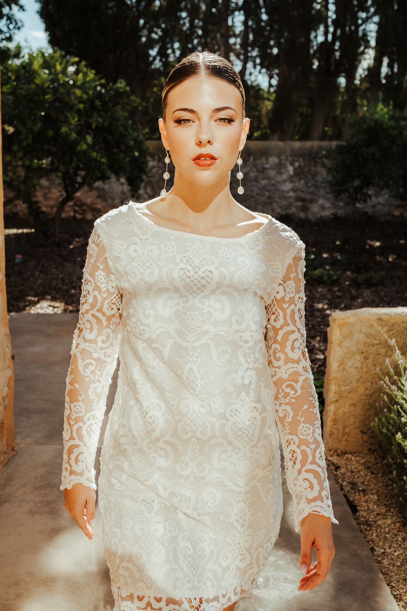 Elegant white lace wedding dress with long sleeves and intricate floral pattern photographed in bright sunlight.
