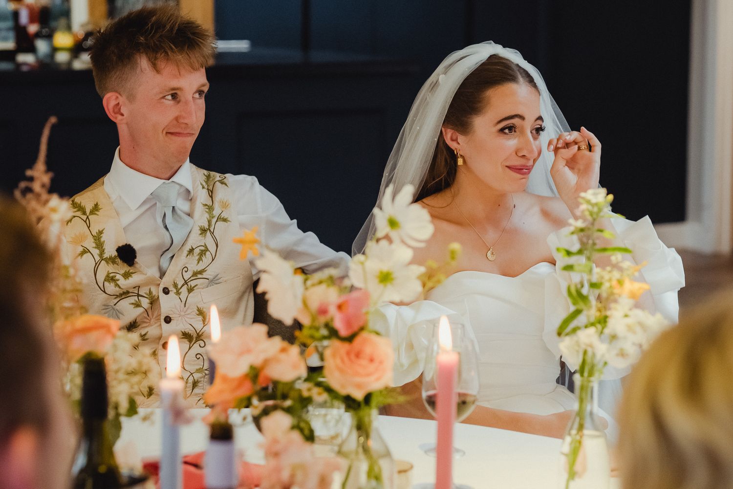 A joyful wedding reception scene with a couple seated at a decorated table with floral centerpieces and candles.