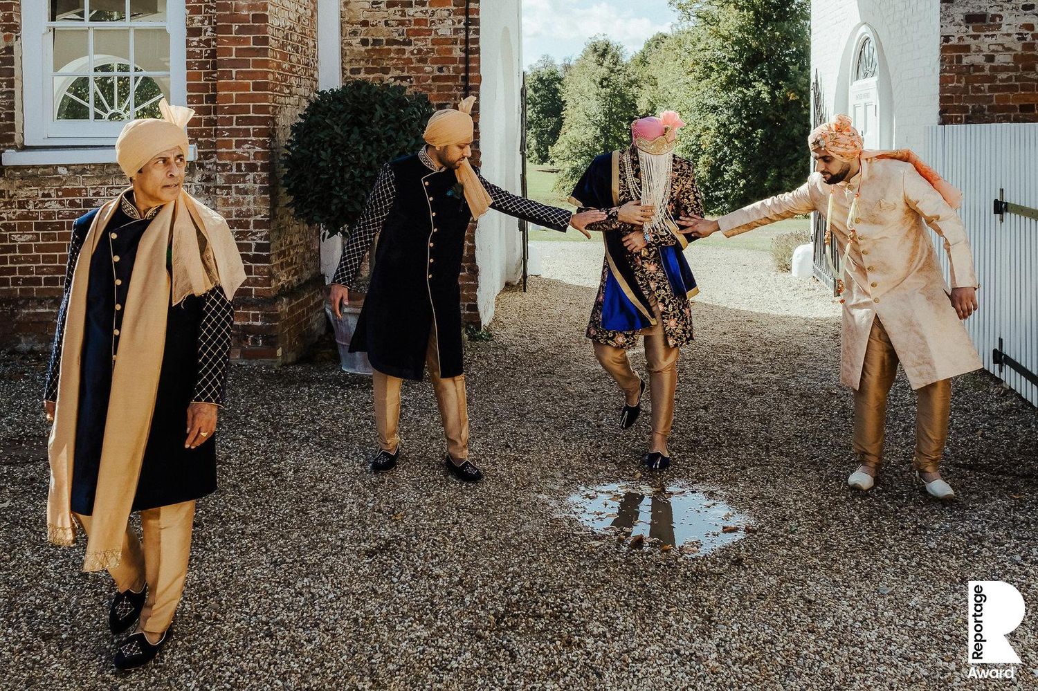 A group of people in stylish coats dance joyfully on gravel outside a brick building.
