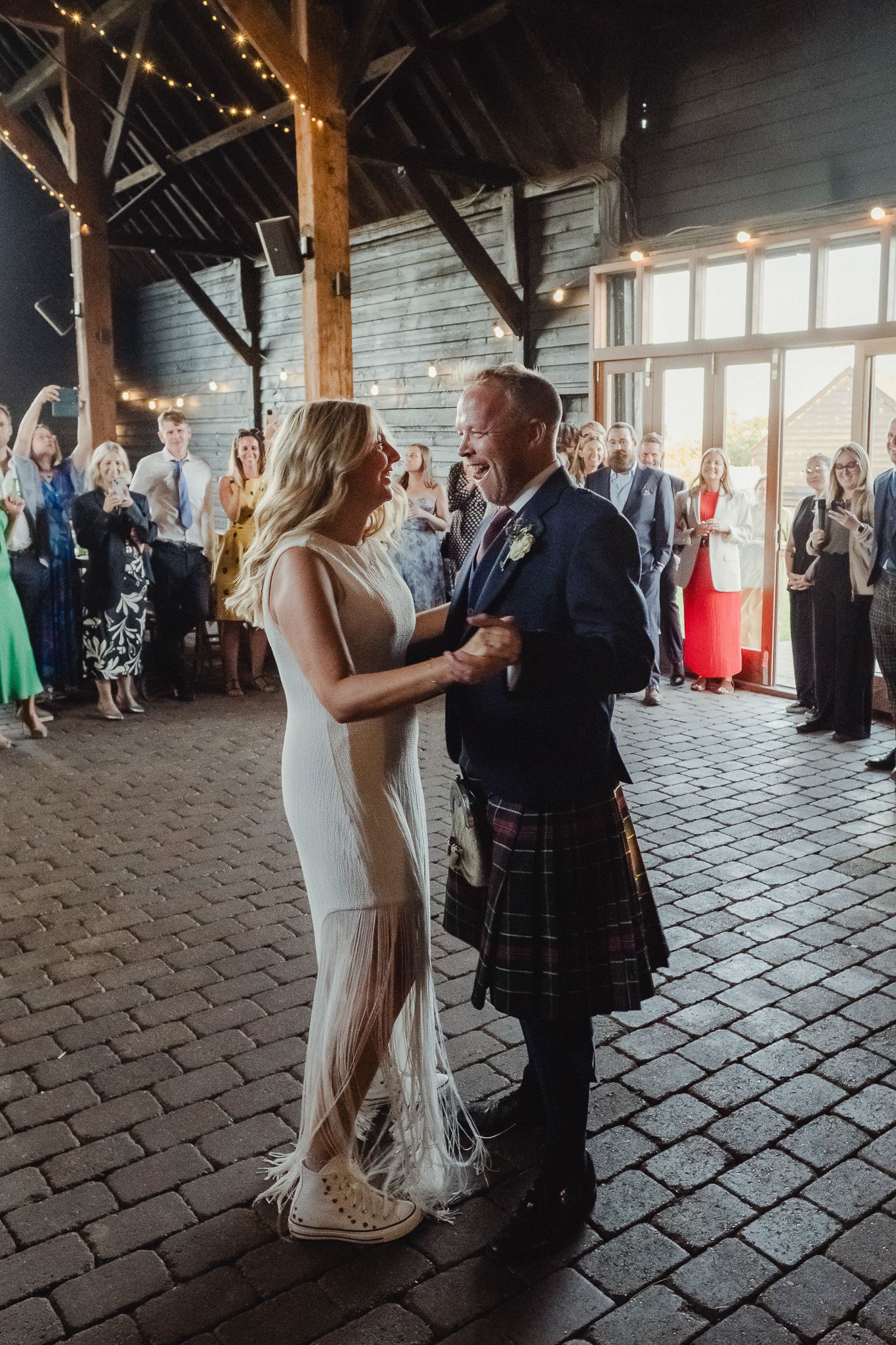 A couple shares their first dance as newlyweds in a rustic barn venue with exposed wooden beams and stone floor.