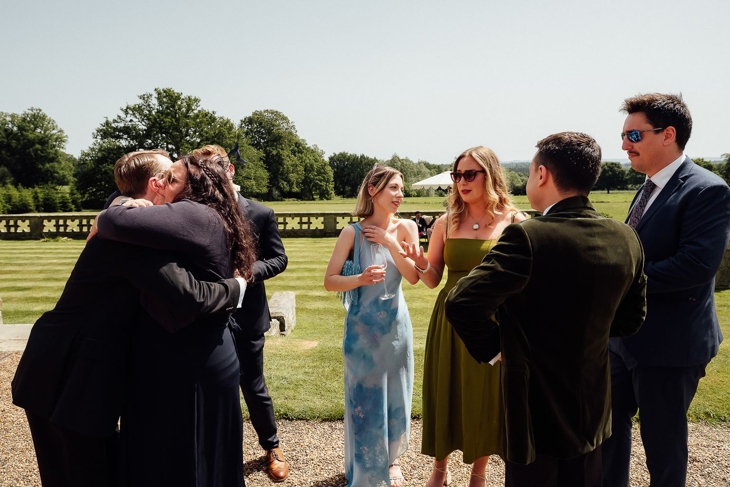 Guests mingling outdoors at a wedding reception on a sunny day, with couples embracing and chatting on a gravel path.