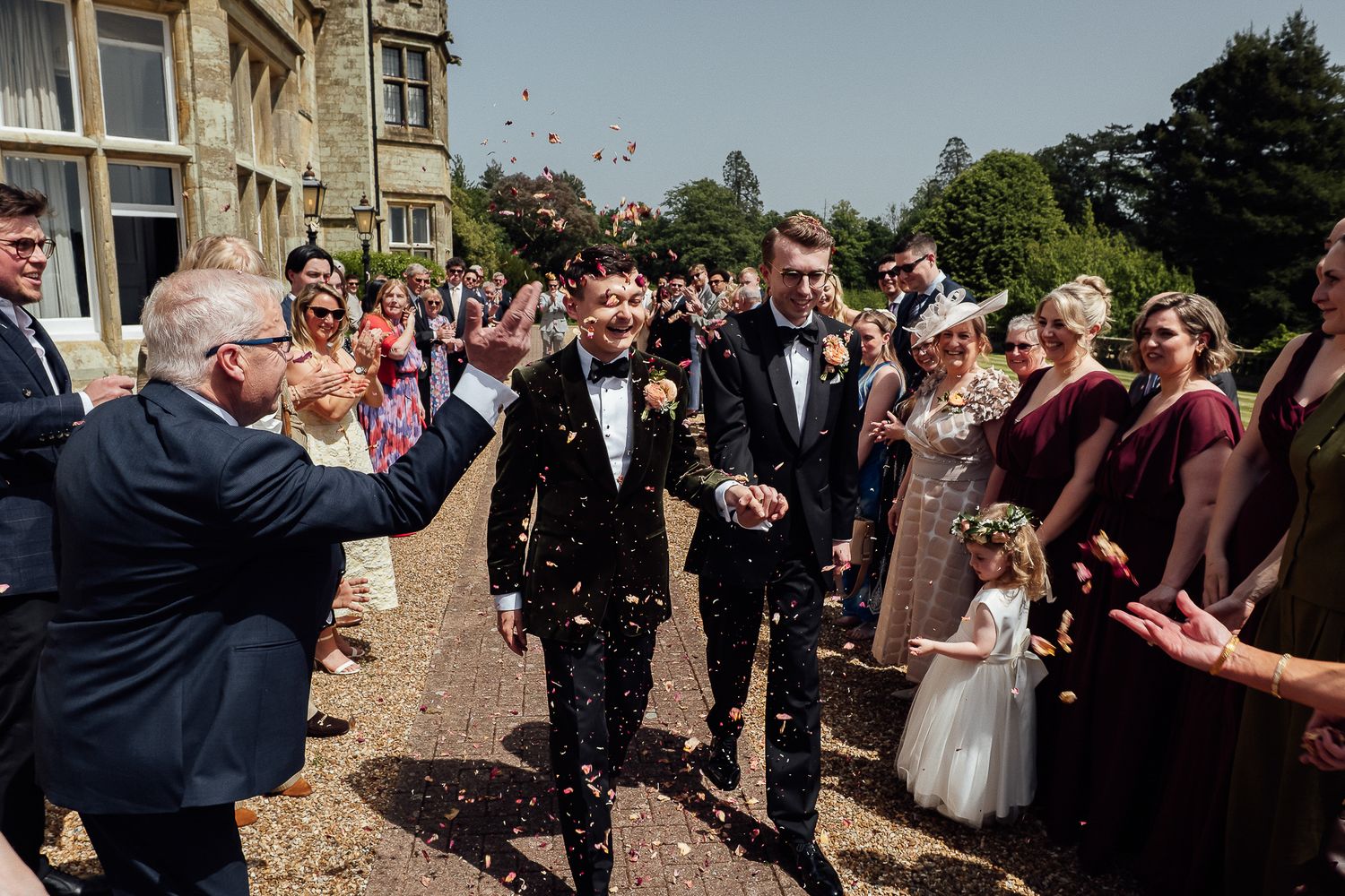 Two grooms in tuxedos walk through a confetti shower surrounded by smiling wedding guests outside a grand stone manor.