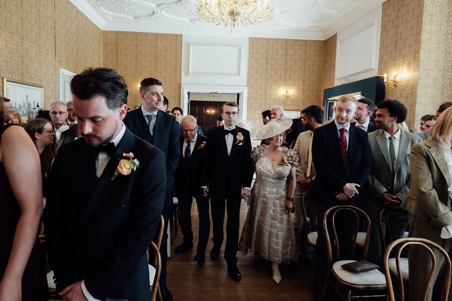 Elegant wedding ceremony procession in a grand room with guests in formal attire, including a woman in a decorative hat.