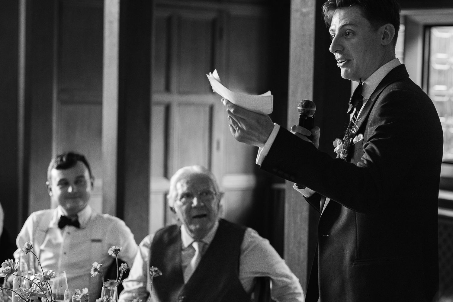 Black and white photo of a man giving a wedding speech while guests listen attentively at a formal reception.
