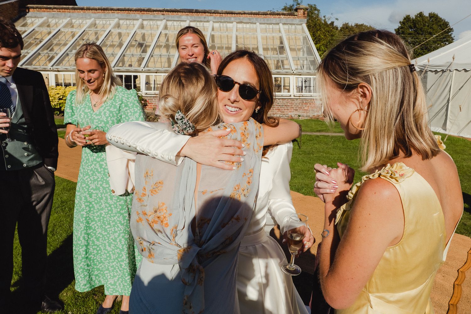 A group of guests enjoying drinks and conversation at an outdoor garden party near a greenhouse on a sunny day.