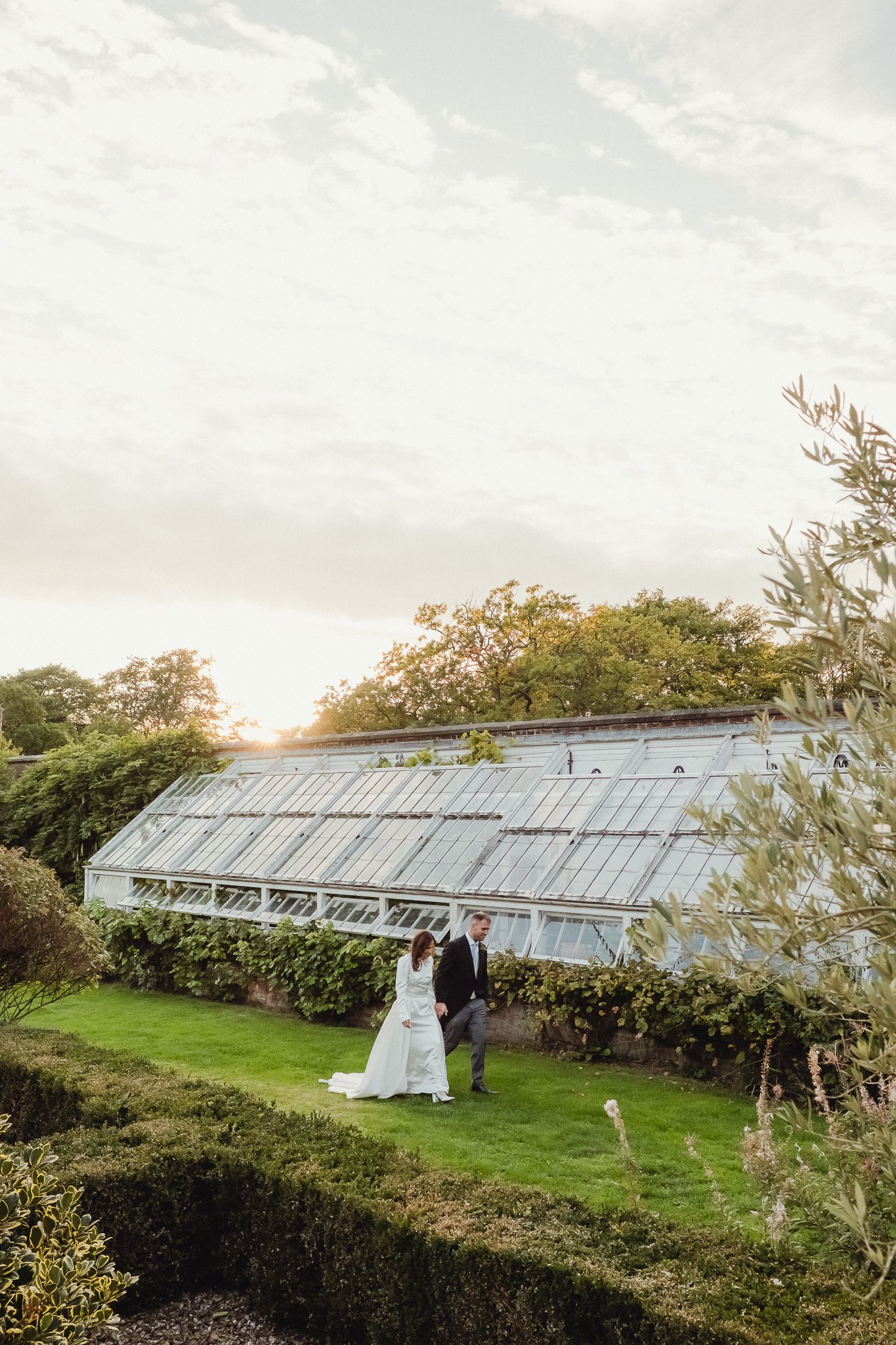 A couple poses in front of a long rustic greenhouse structure during a wedding photo session at sunset.