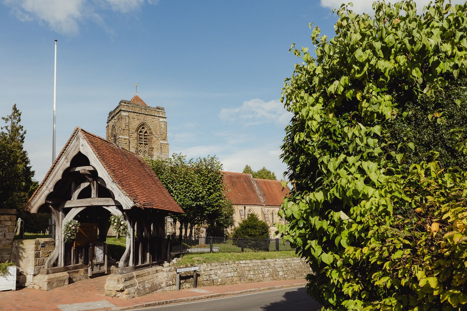 A panoramic view of a historic church with a bell tower and lych gate surrounded by lush green trees in summer.