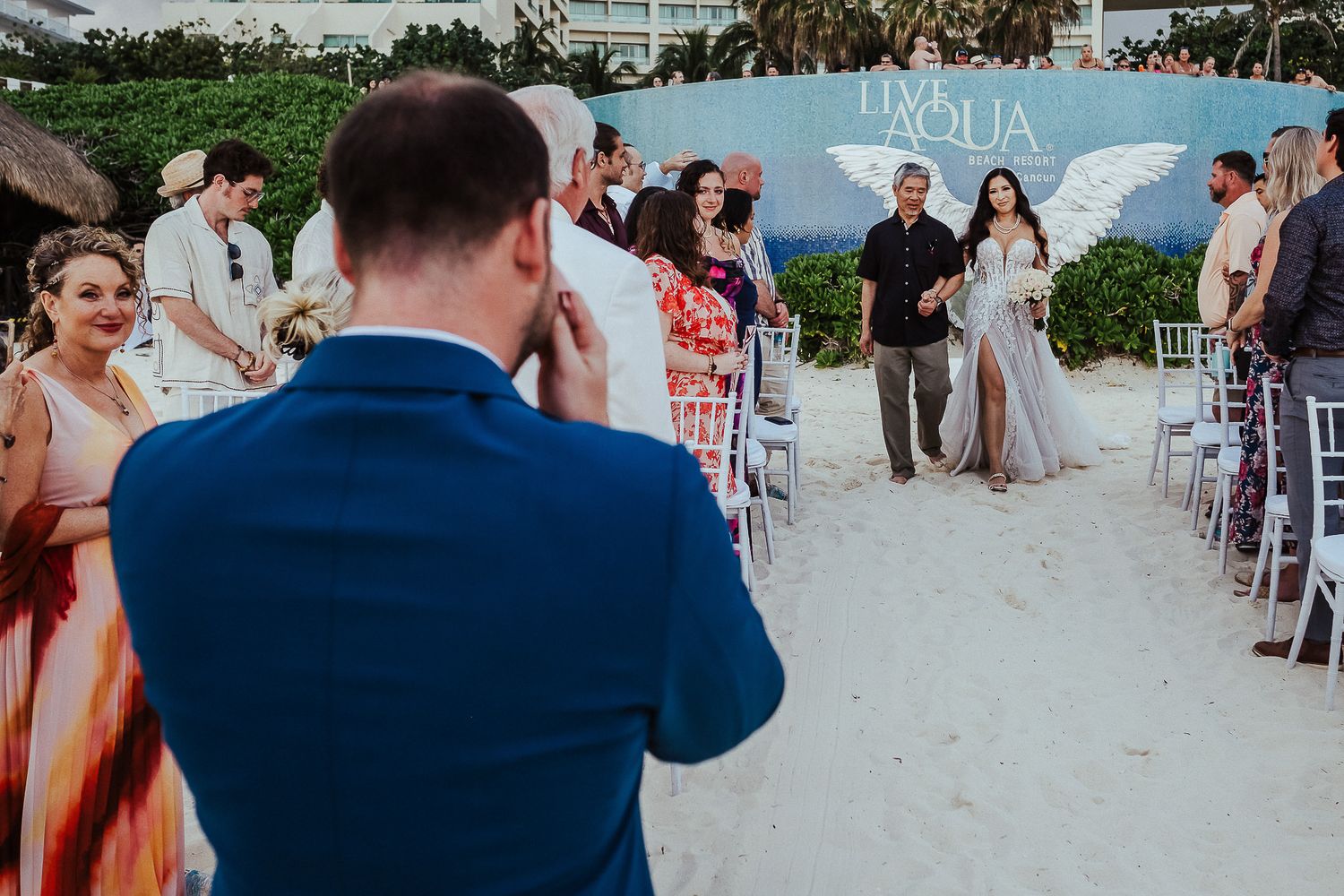 Wedding ceremony guests stand in rows on a beach with palm trees and ocean views in the background.