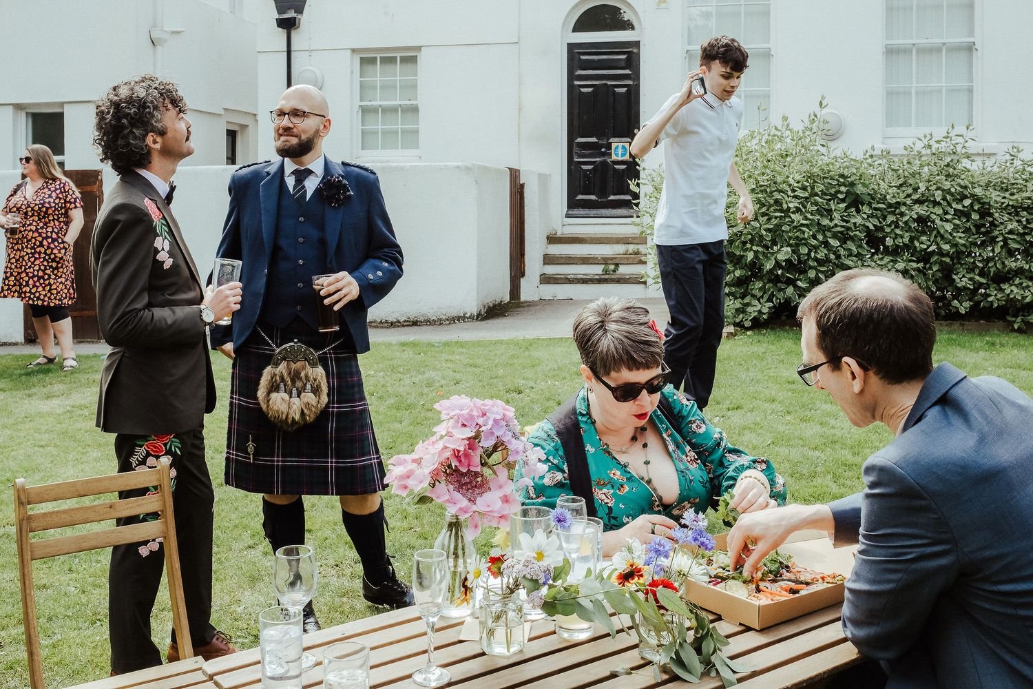Wedding guests gather in a garden, a man in a kilt chatting while others sit at a table of colorful flower arrangements.
