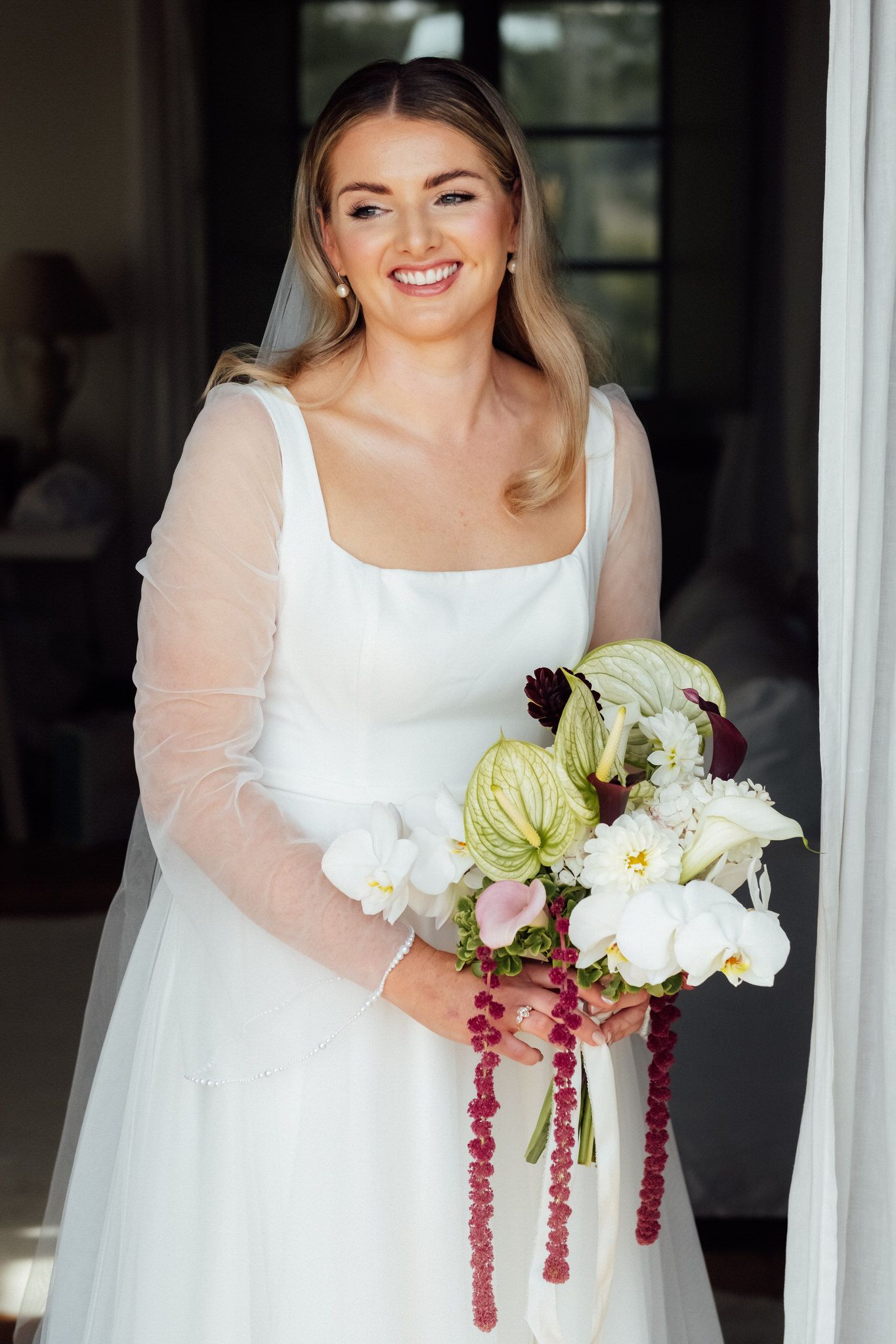 Smiling bride in a white square-neck gown with sheer sleeves and veil holds a bouquet of white anthuriums, green blooms and red amaranthus.