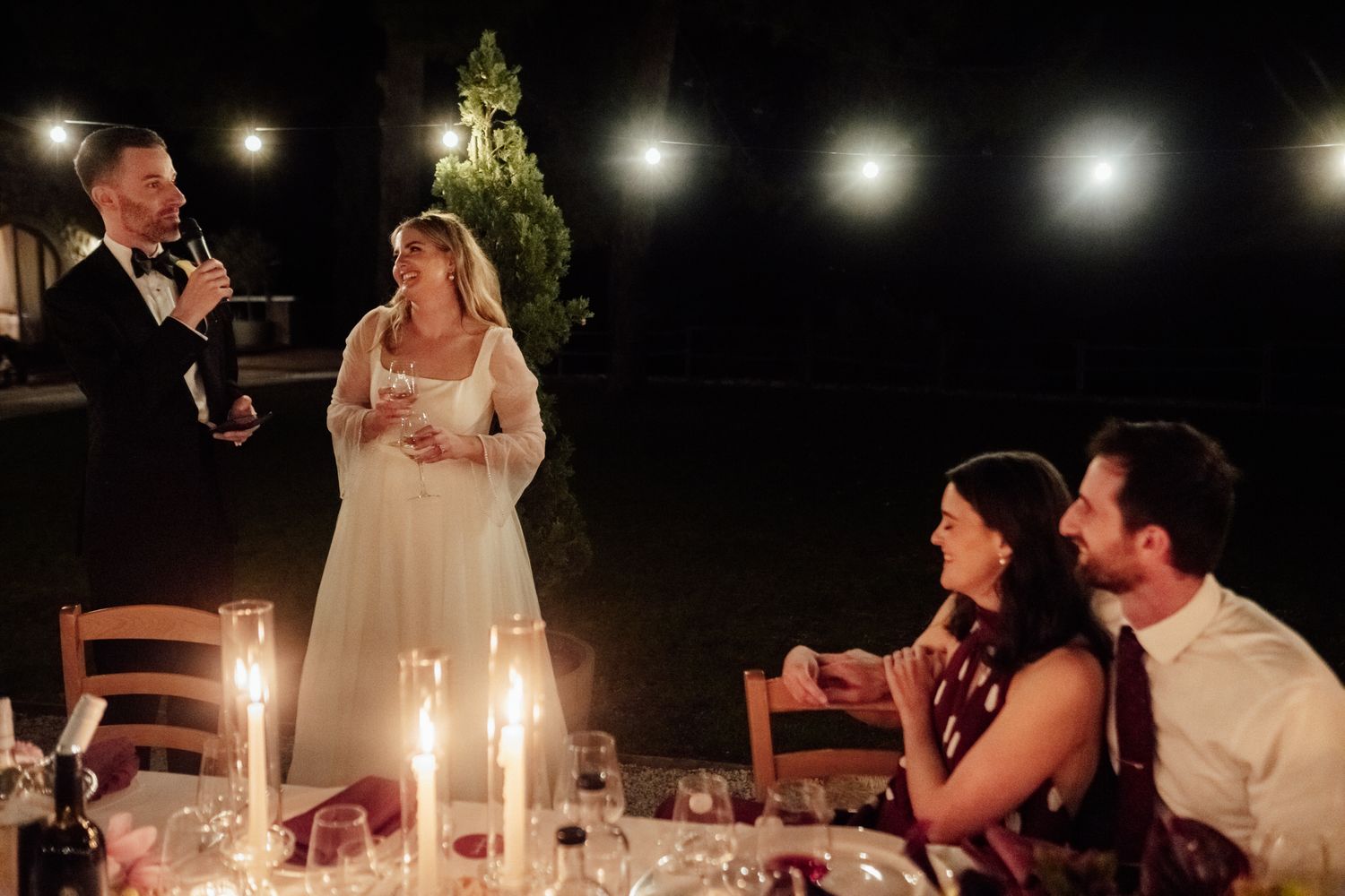 Bride in a white gown speaks into a microphone during an evening wedding reception while seated guests smile at candlelit tables.