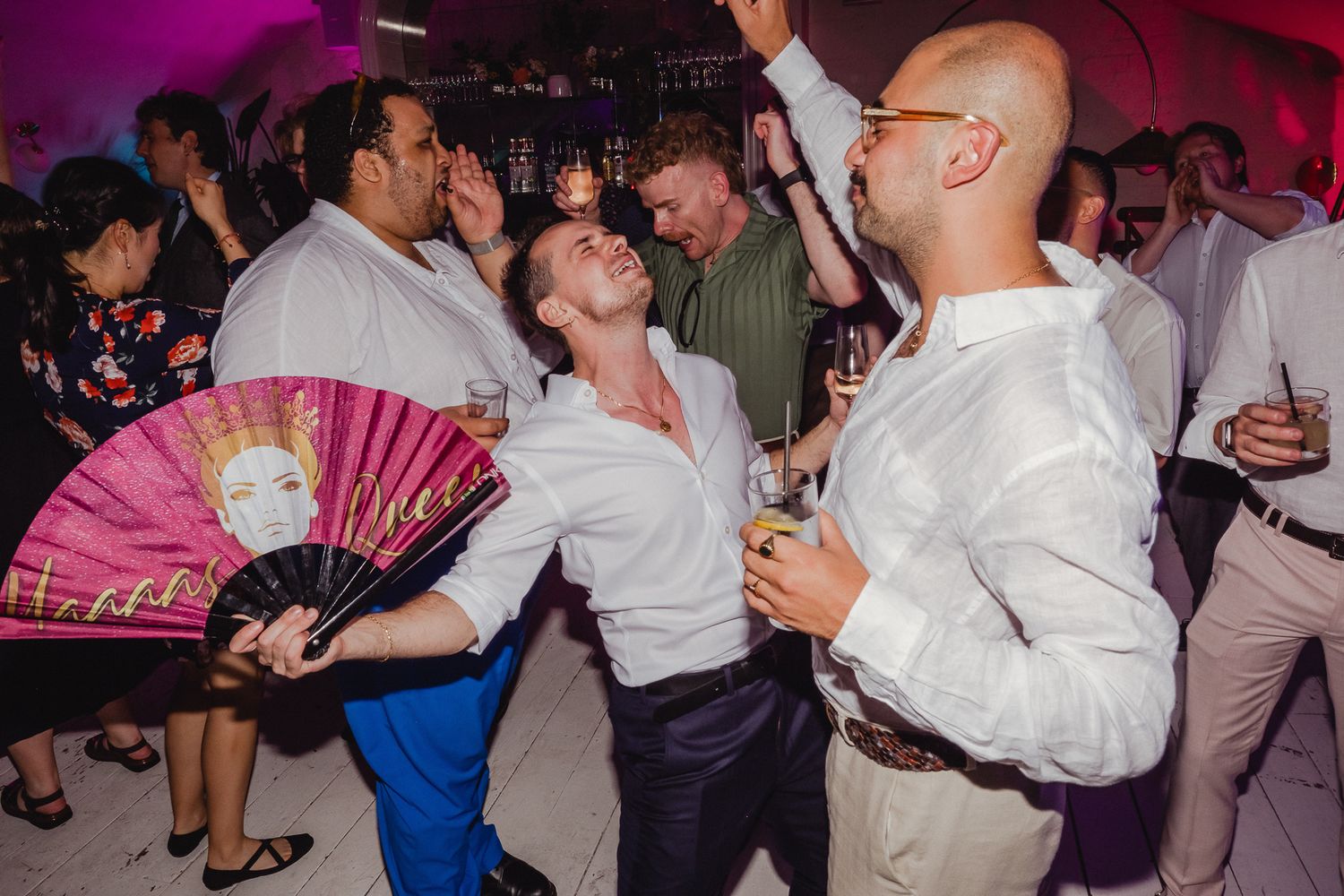 People dancing energetically at a party with pink lighting while one person holds a decorative fan.