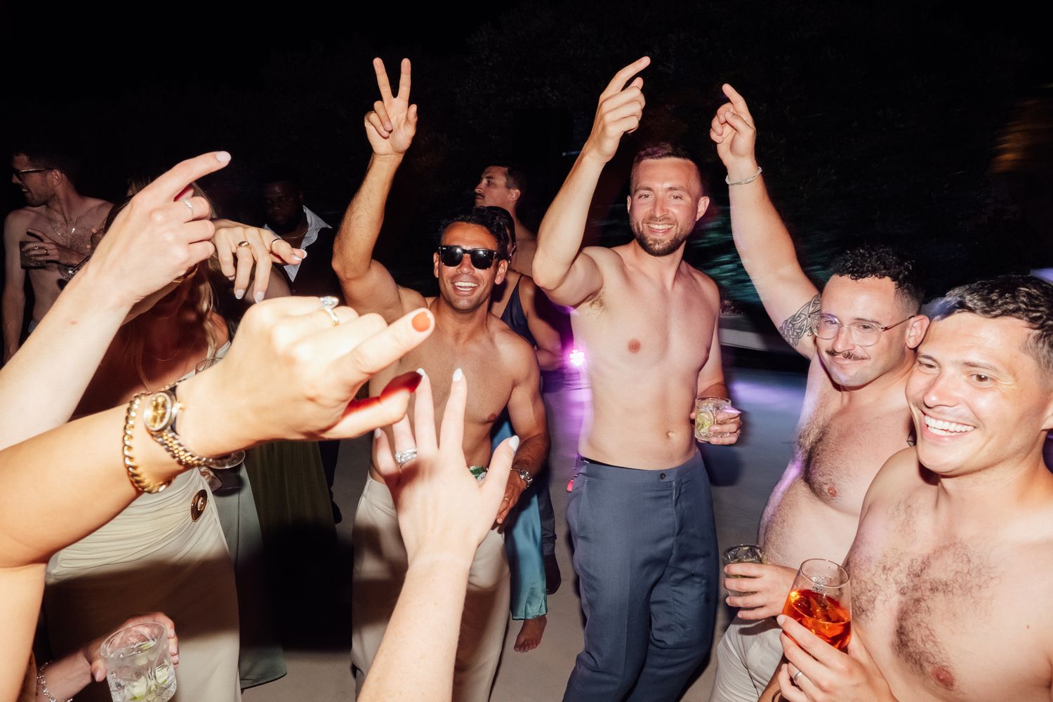 Shirtless men dancing and celebrating at an outdoor nighttime beach party with colorful lights.