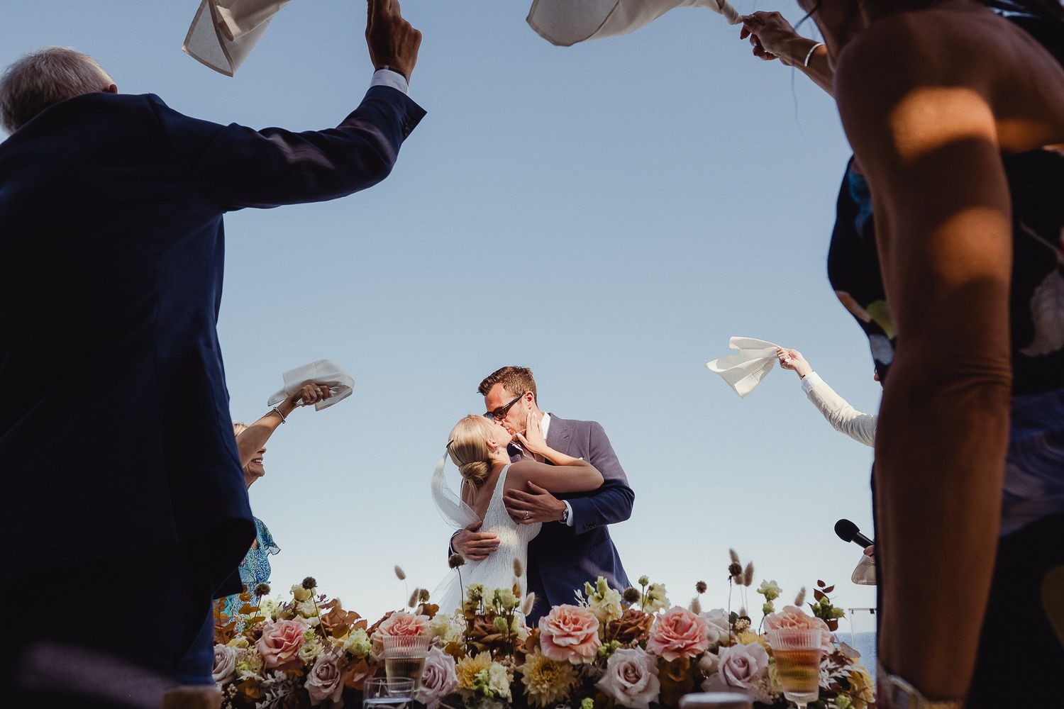 A low-angle view captures people throwing paper airplanes against a bright blue sky with trees in the background.
