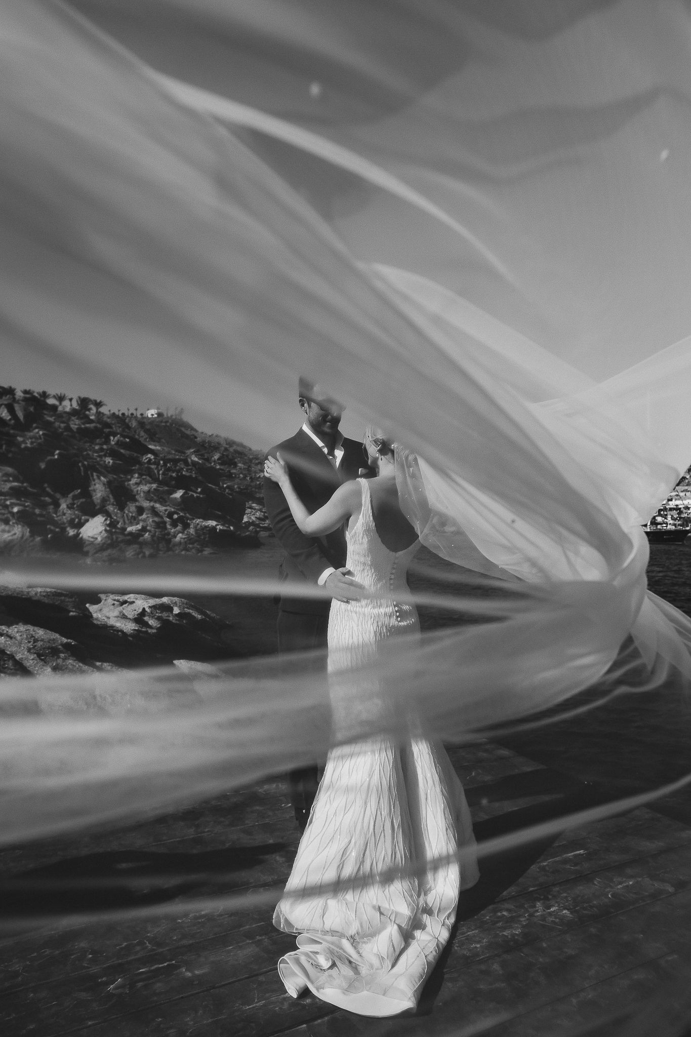 A flowing white wedding dress veil billows dramatically against a coastal landscape in black and white.
