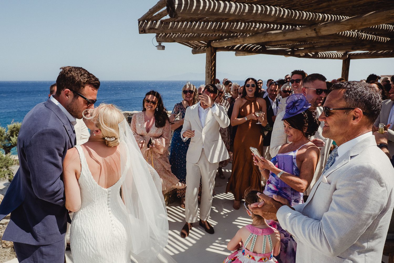 Wedding guests seated on a beach under a wooden pergola during an outdoor ceremony by the ocean.