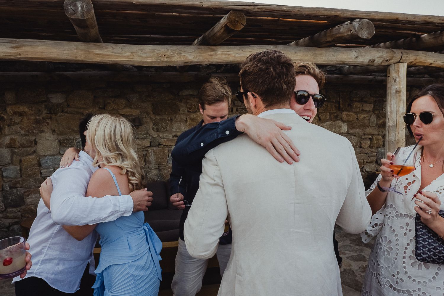 Group of people sharing emotional embraces and hugs at an outdoor gathering near a stone building.