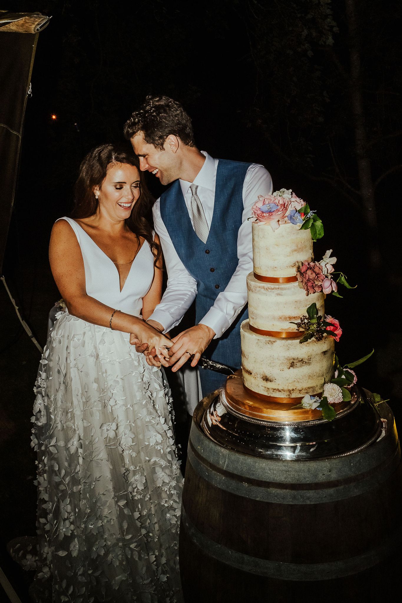 Elegant three-tiered wedding cake being cut at an evening outdoor reception.