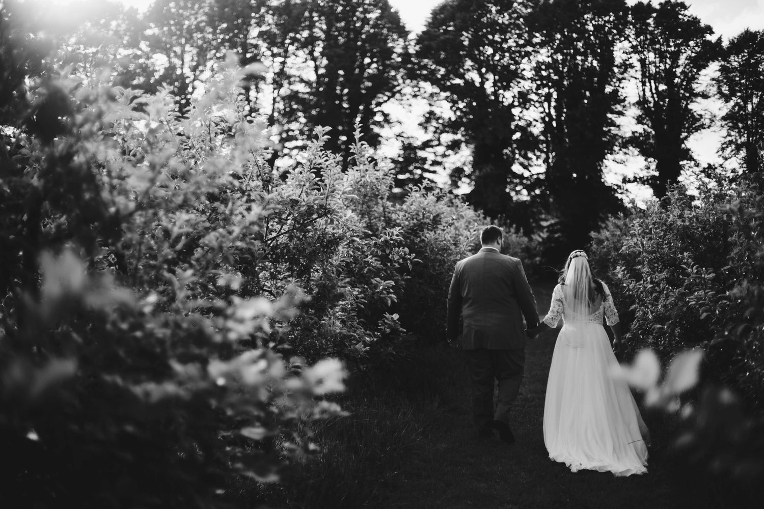 Black and white photo of couple walking through wooded path in wedding attire.