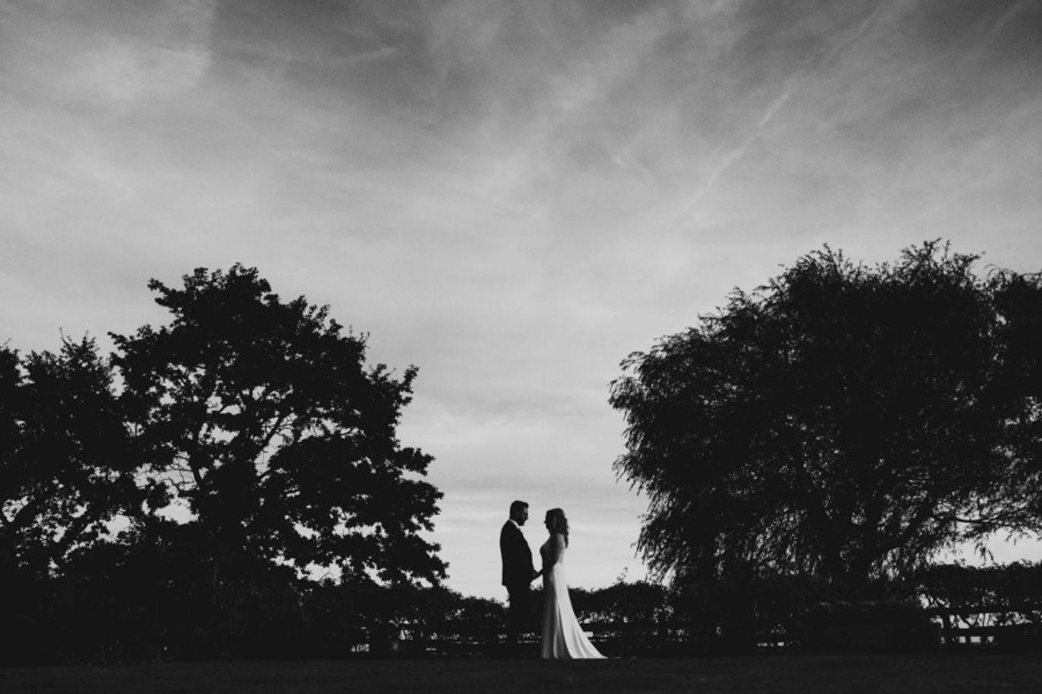 A silhouetted couple stands against a dramatic sky in this black and white wedding portrait.