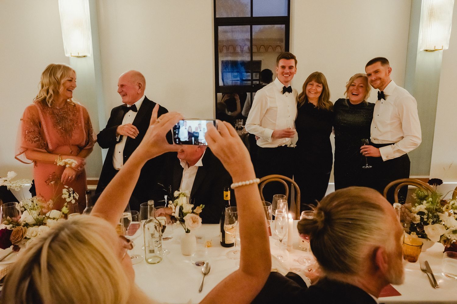 Wedding guests at a reception dinner table raise their glasses in a celebratory toast.