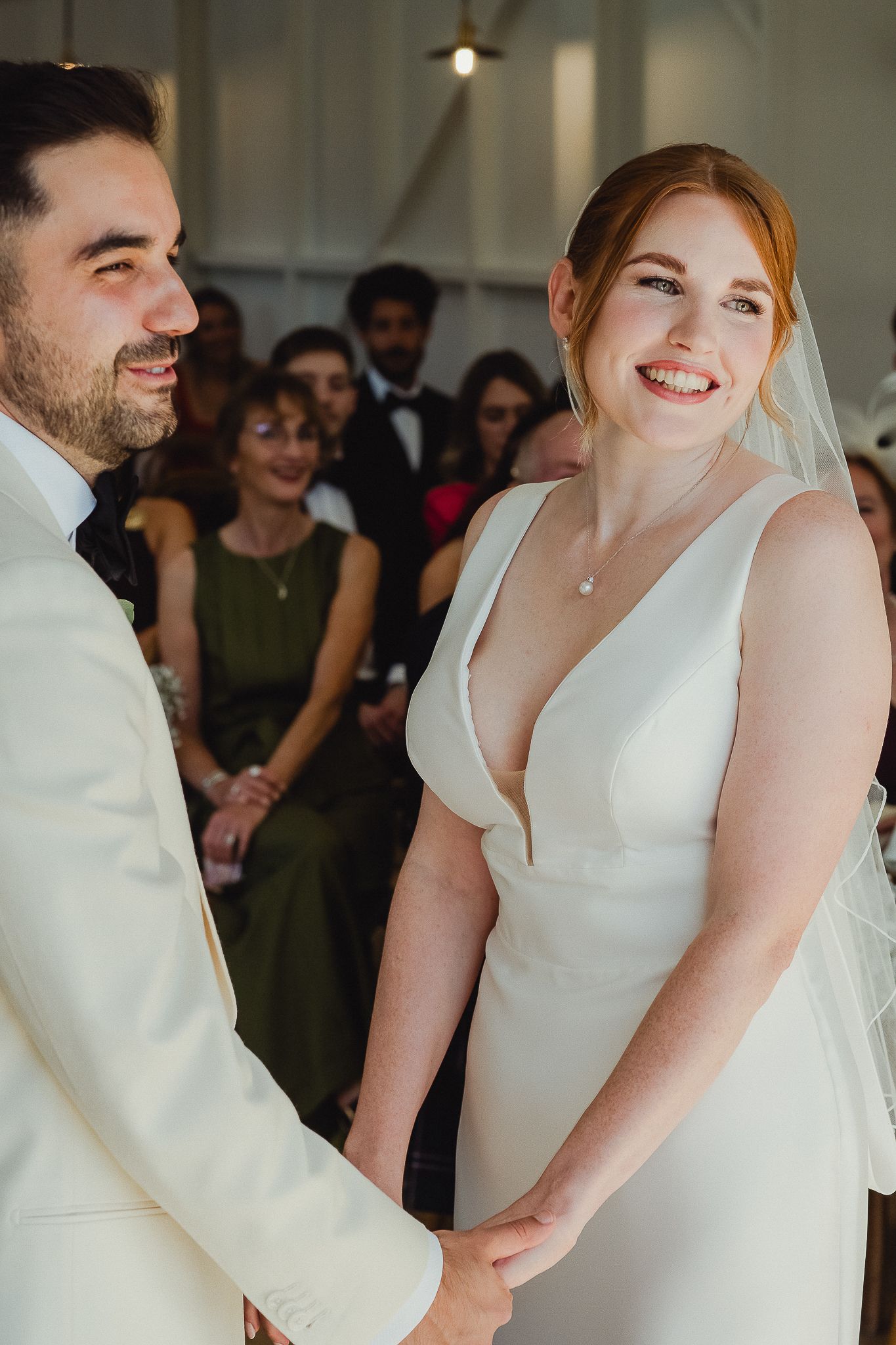 A couple shares a tender moment at their wedding celebration, both wearing white and smiling warmly.
