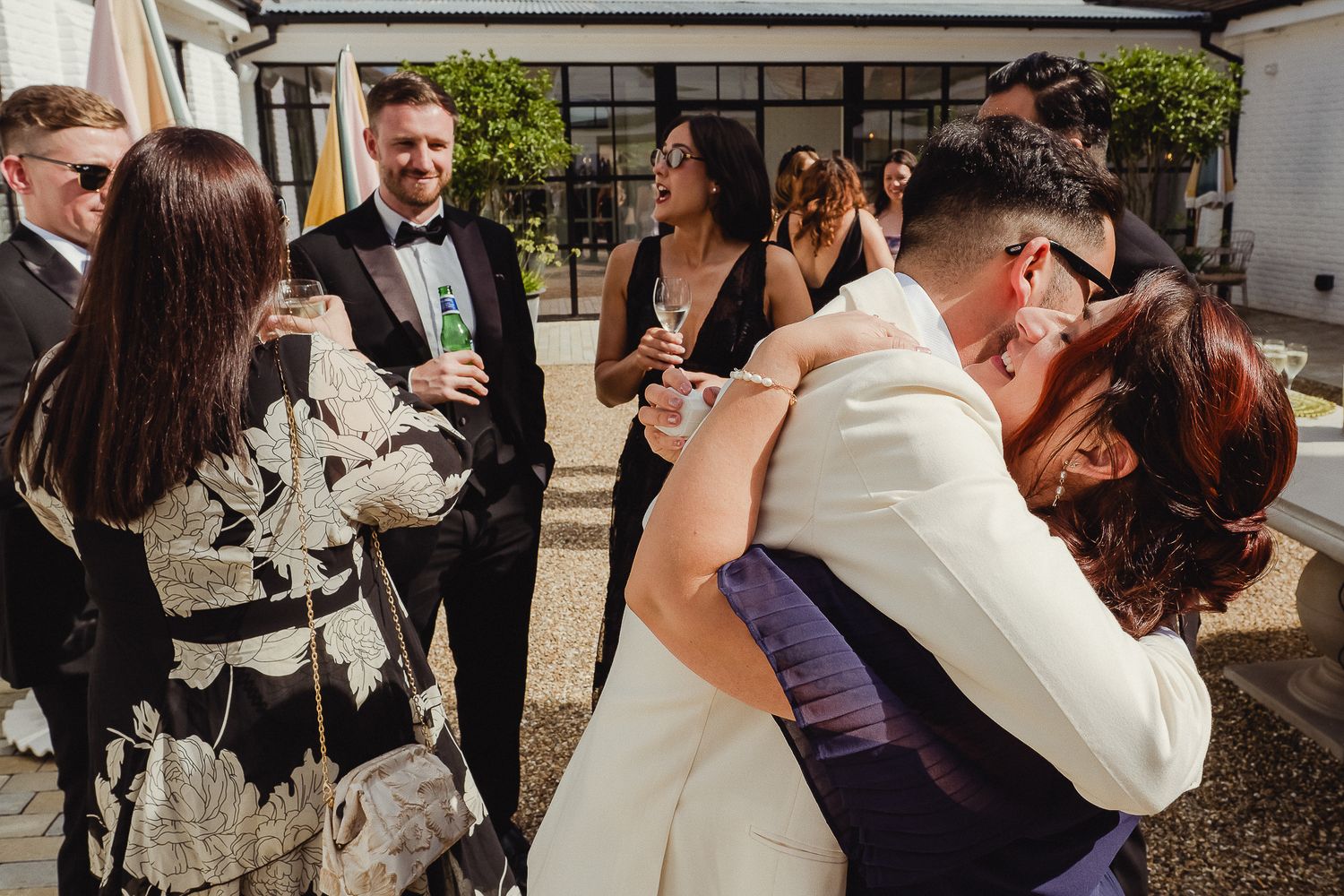 Wedding guests mingle and celebrate during an outdoor reception at a venue with white walls and greenery.