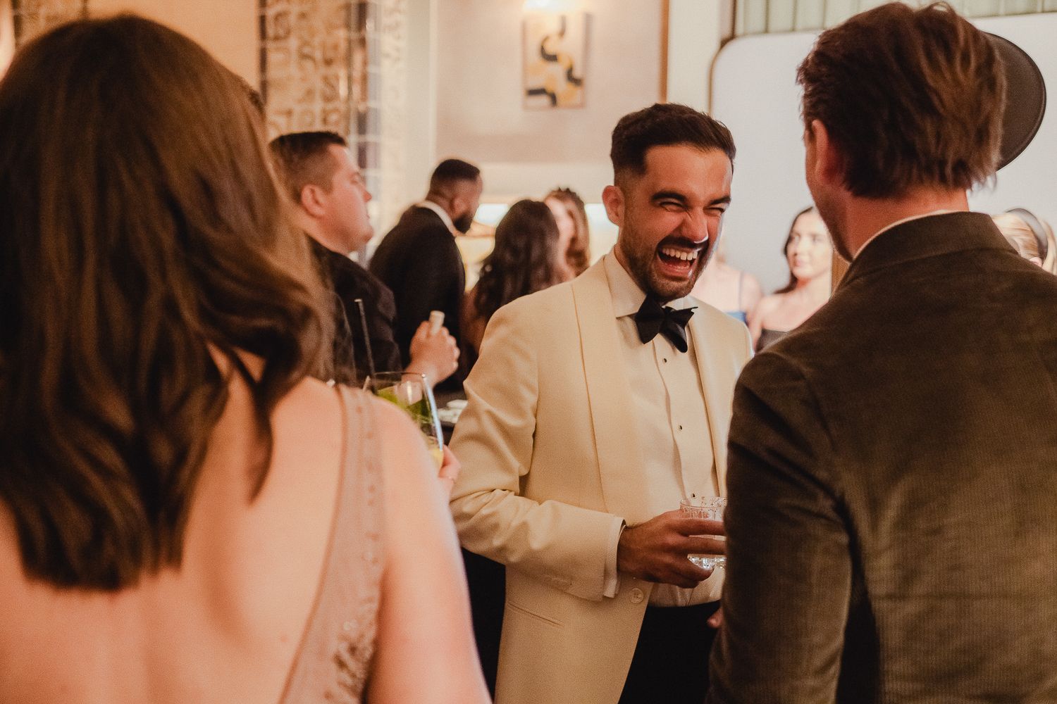 A candid moment at a wedding reception with guests mingling and laughing in a warm-lit indoor venue.