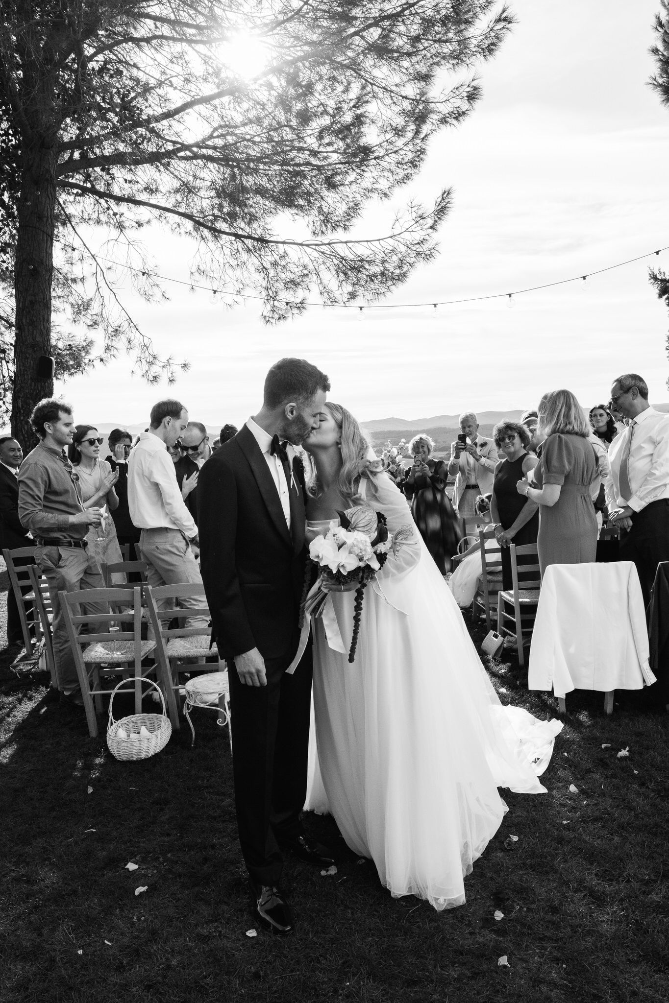 Black and white photo of a bride and groom kissing in an outdoor aisle as wedding guests look on beneath a tree with string lights.