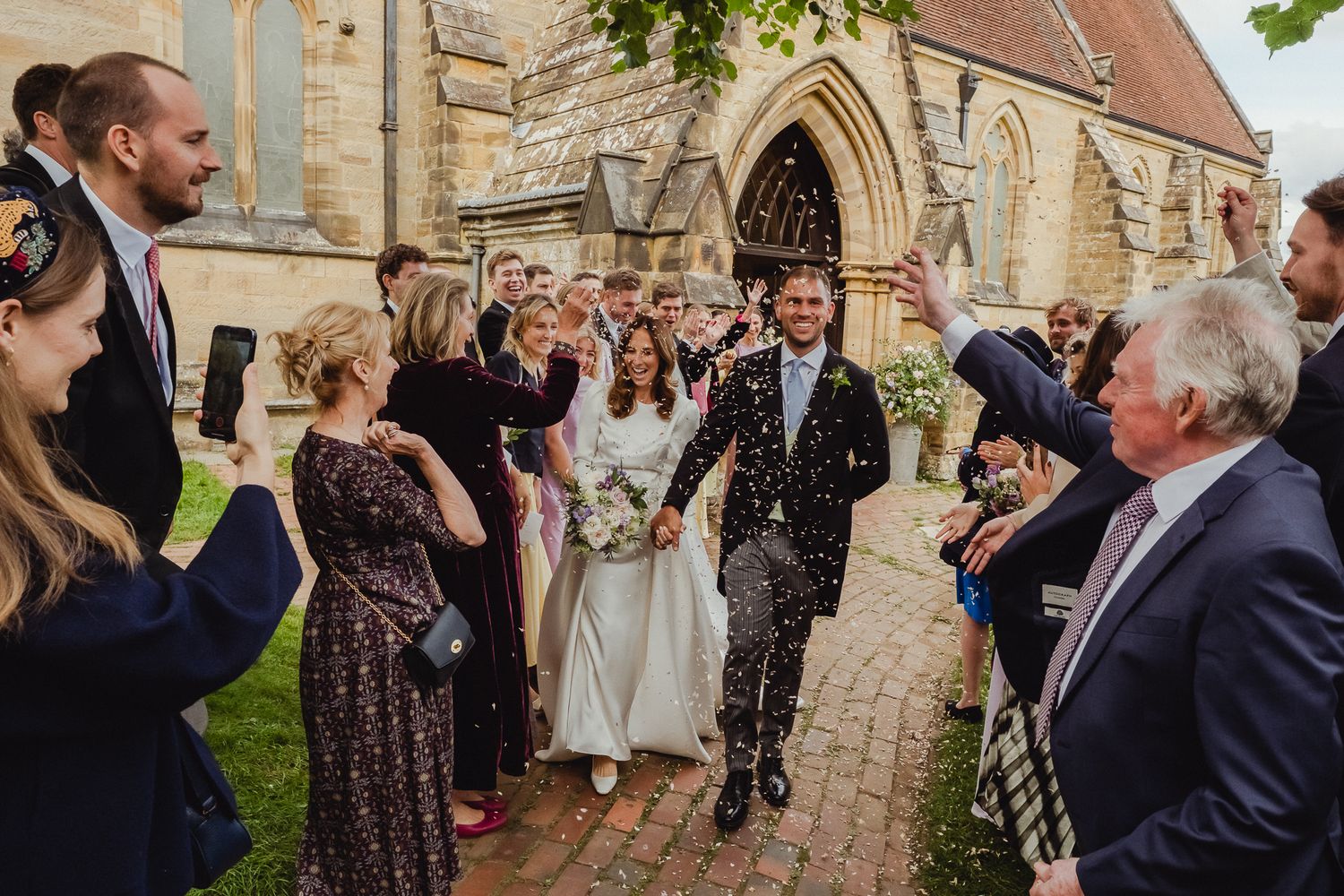 Wedding party celebrates outside historic stone church as guests throw confetti on sunny afternoon.