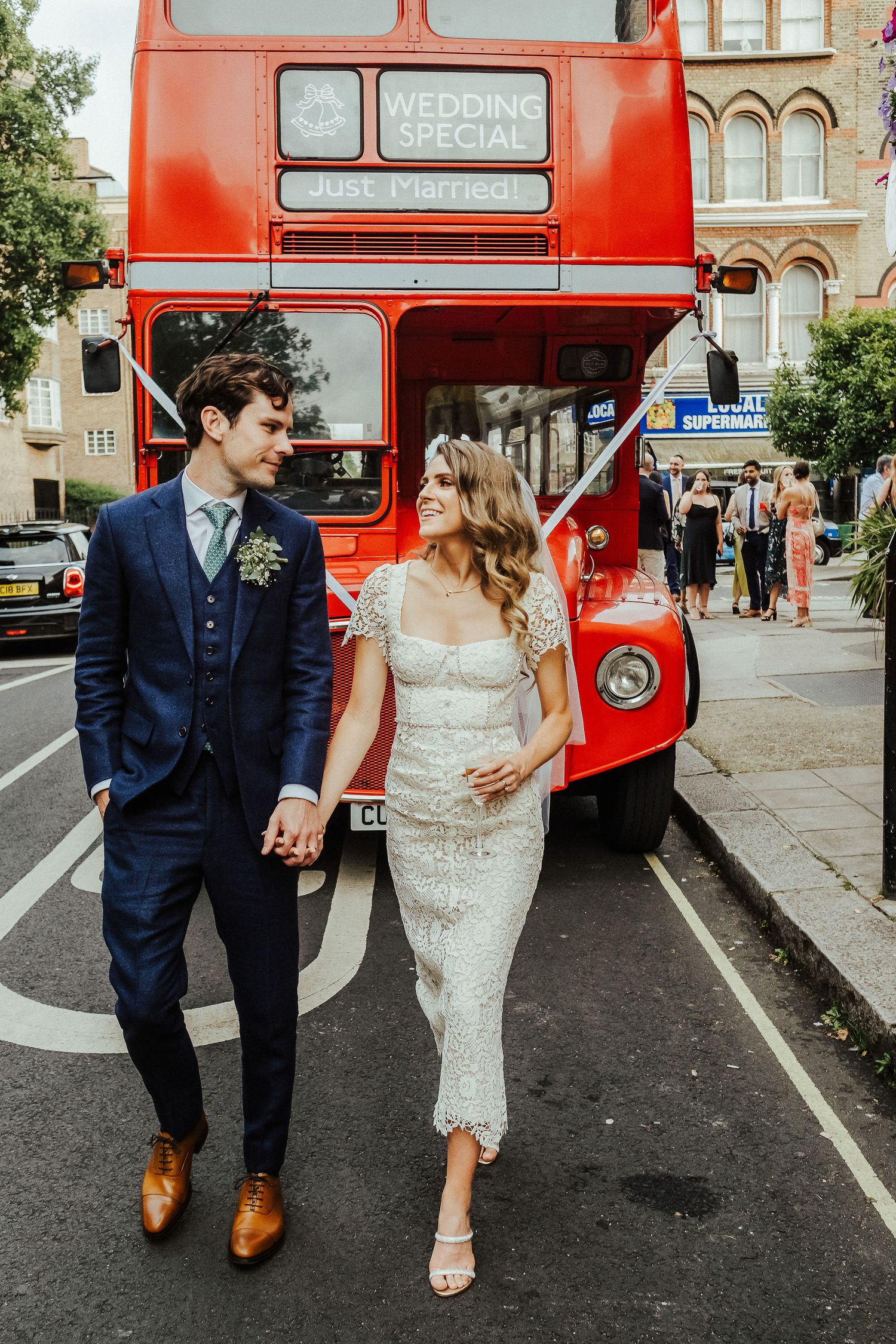 A couple walks hand in hand past a classic red London double decker bus during their wedding celebration.
