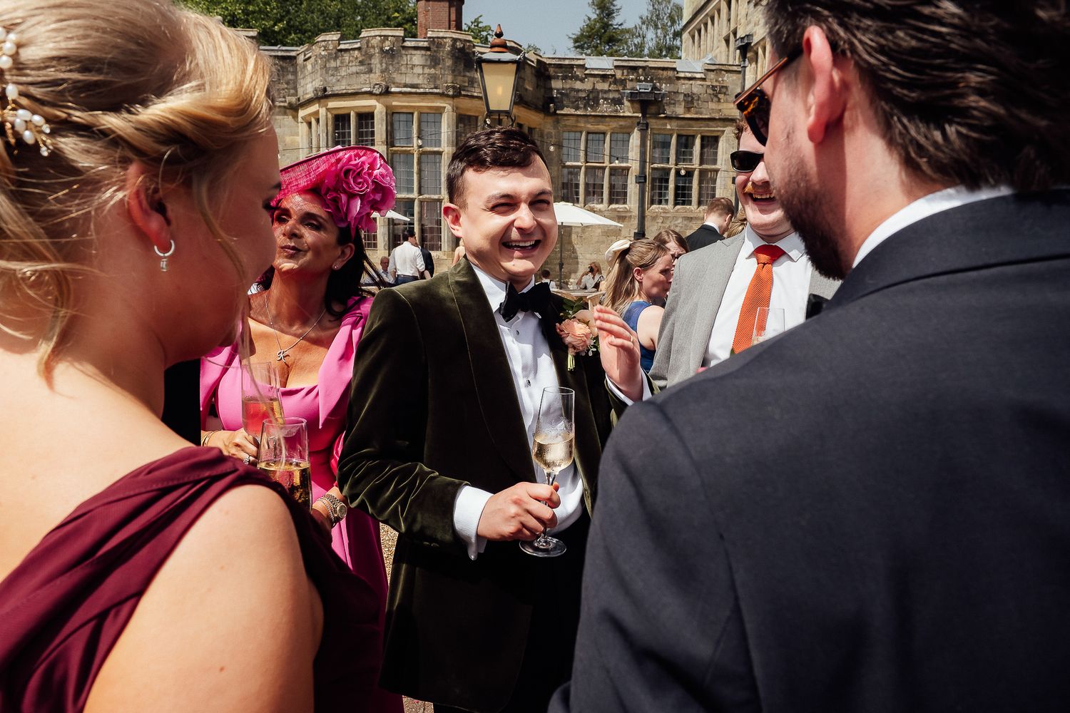 Elegantly dressed wedding guests socializing outdoors, including a woman in pink fascinator and man in dark suit holding champagne.