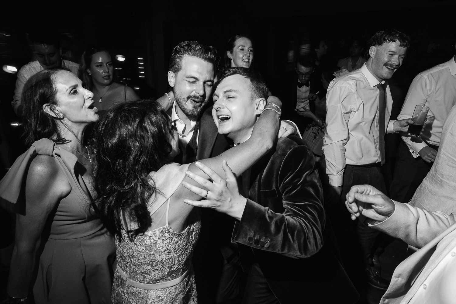Black and white wedding reception photo of guests joyfully dancing and embracing on the dance floor.