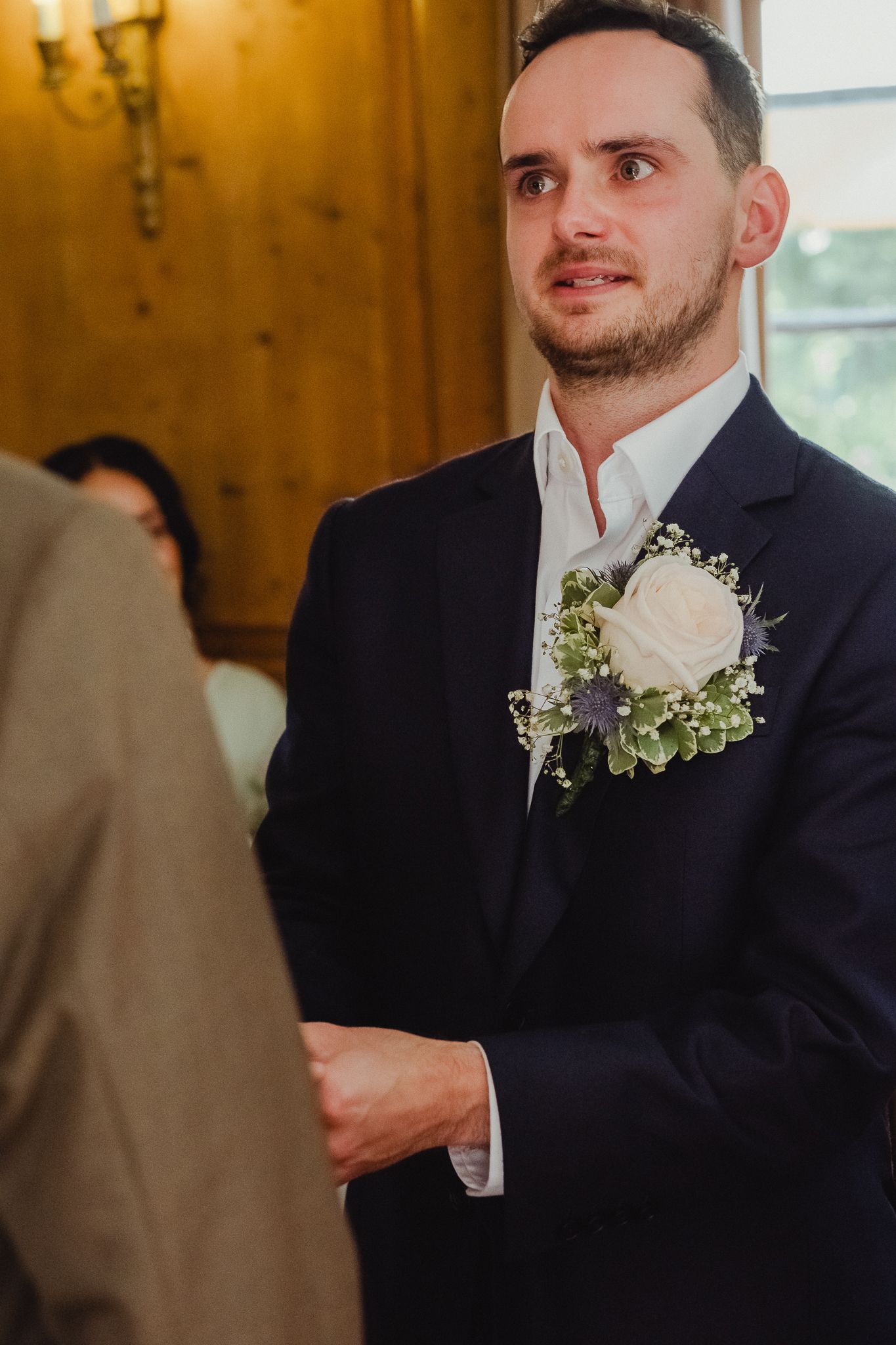 Groom in navy suit wearing white rose and purple floral boutonniere smiles during wedding ceremony.