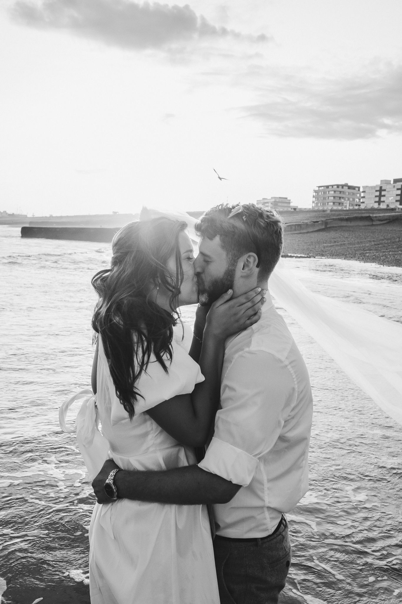 A romantic black and white photo series of a couple embracing on a beach with waves and buildings in the background.