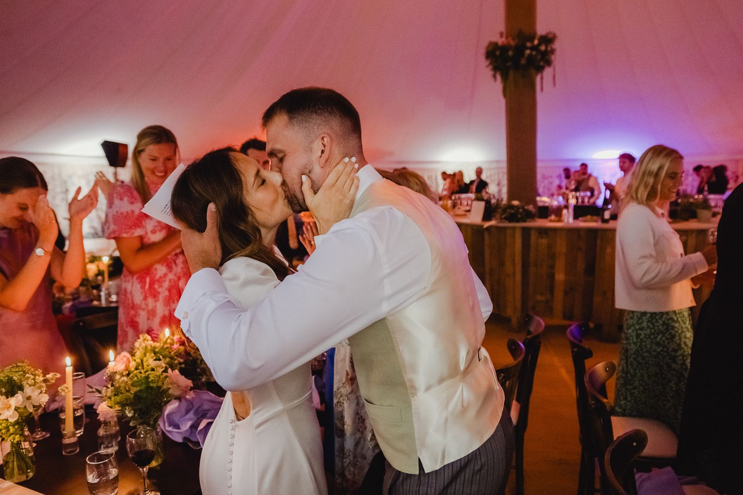 A couple shares a romantic kiss on the dance floor at a wedding reception under pink and purple lighting.
