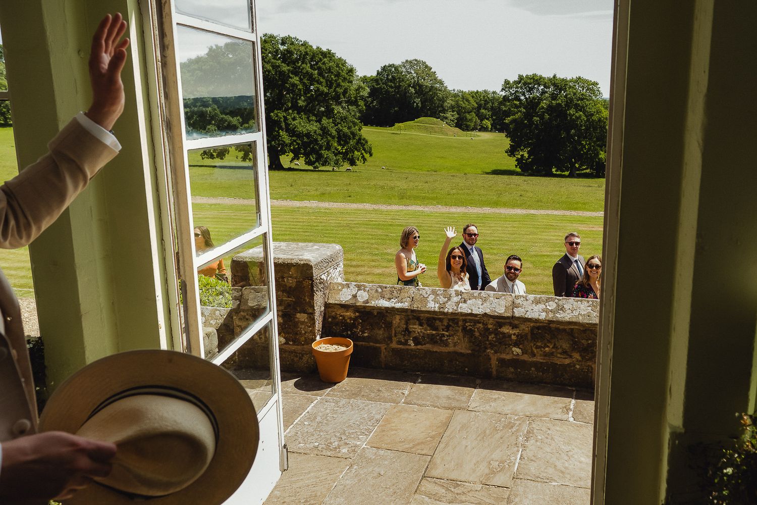 View through an open window overlooking people gathered on a stone patio with rolling green hills in the background.