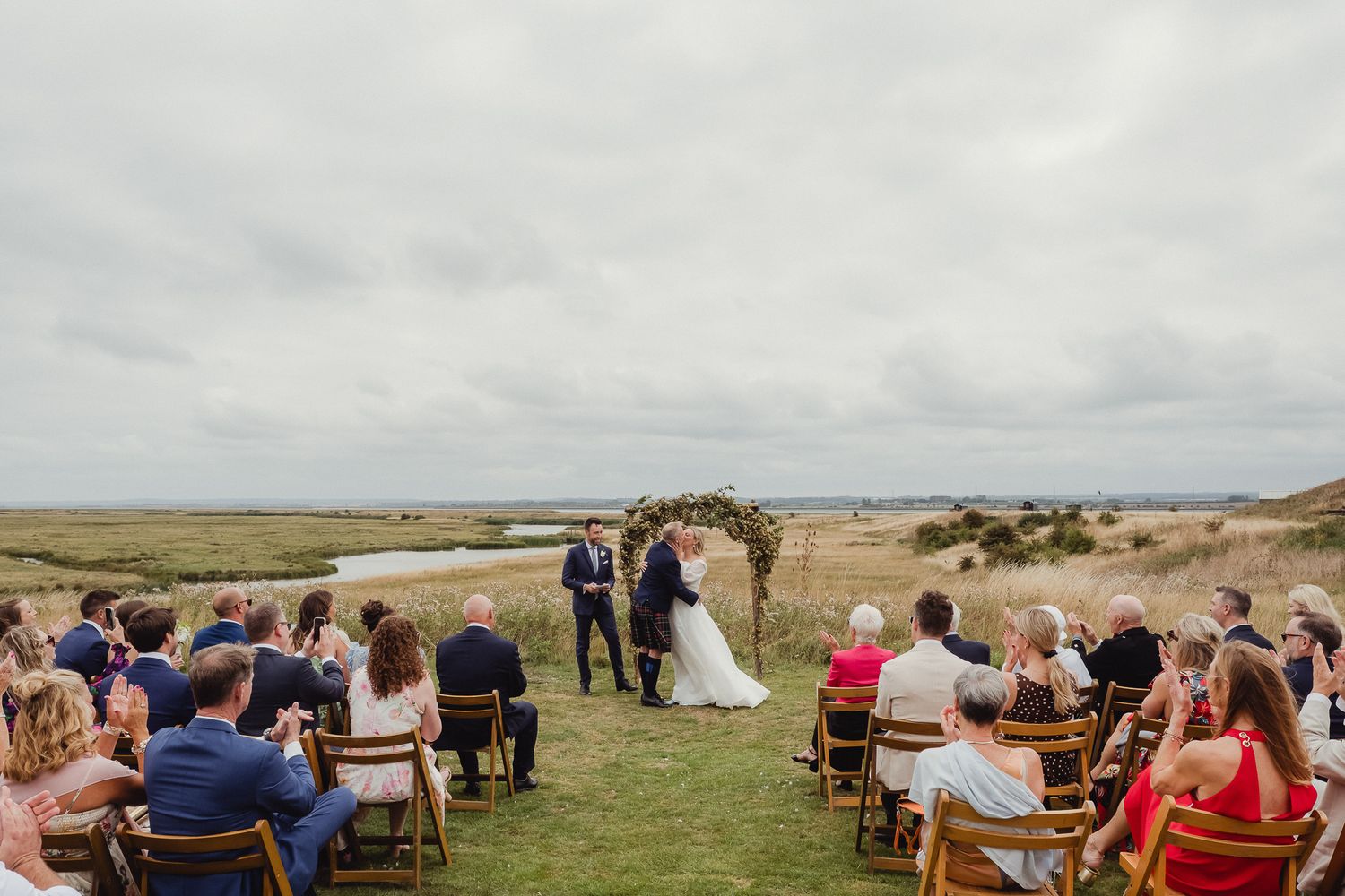 A panoramic view of an outdoor wedding ceremony set against a dramatic coastal landscape with guests seated in rows.