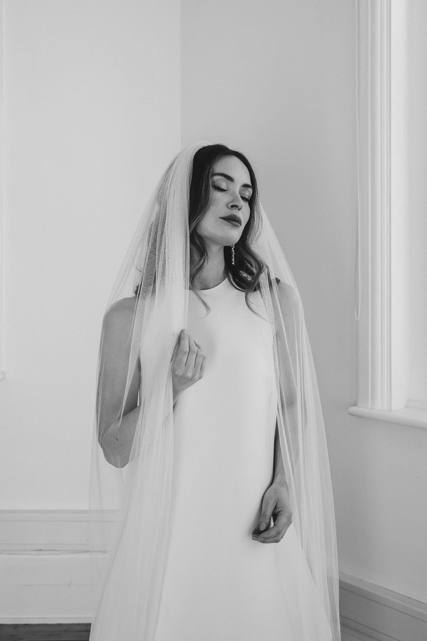 A bride in a minimalist white dress and long veil poses elegantly in a bright white room for a series of artistic photos.