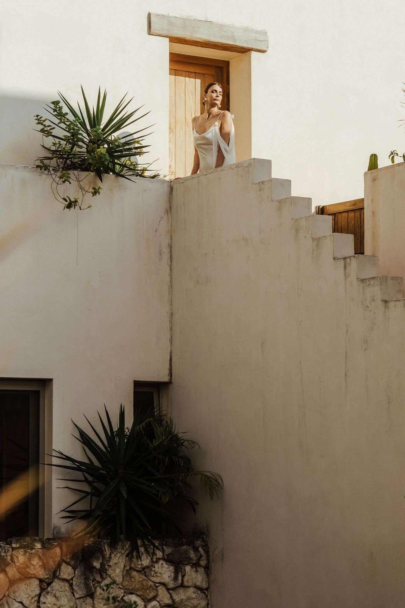 White stucco walls with geometric steps create an architectural pattern against a bright sky, adorned with tropical plants.