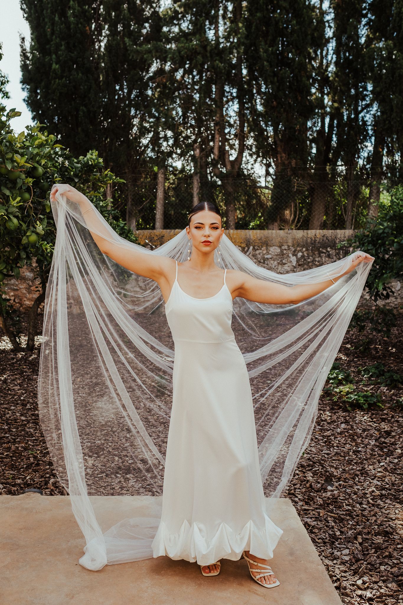 Bride in a flowing white dress holds up a delicate veil in an outdoor garden setting.
