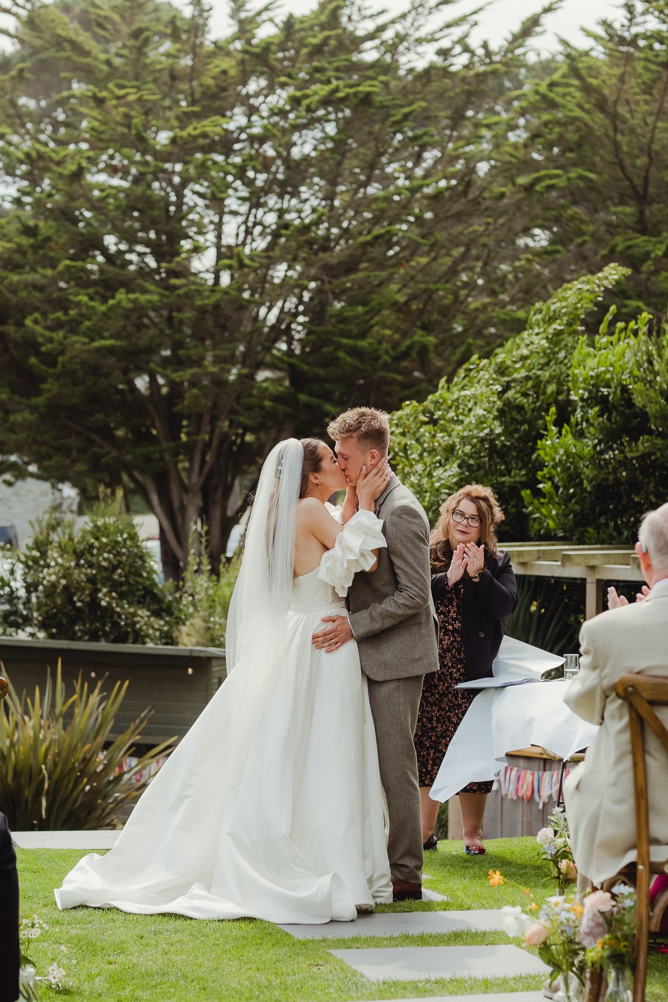 Newlywed couple shares their first kiss during an outdoor garden wedding ceremony as guests celebrate joyfully.