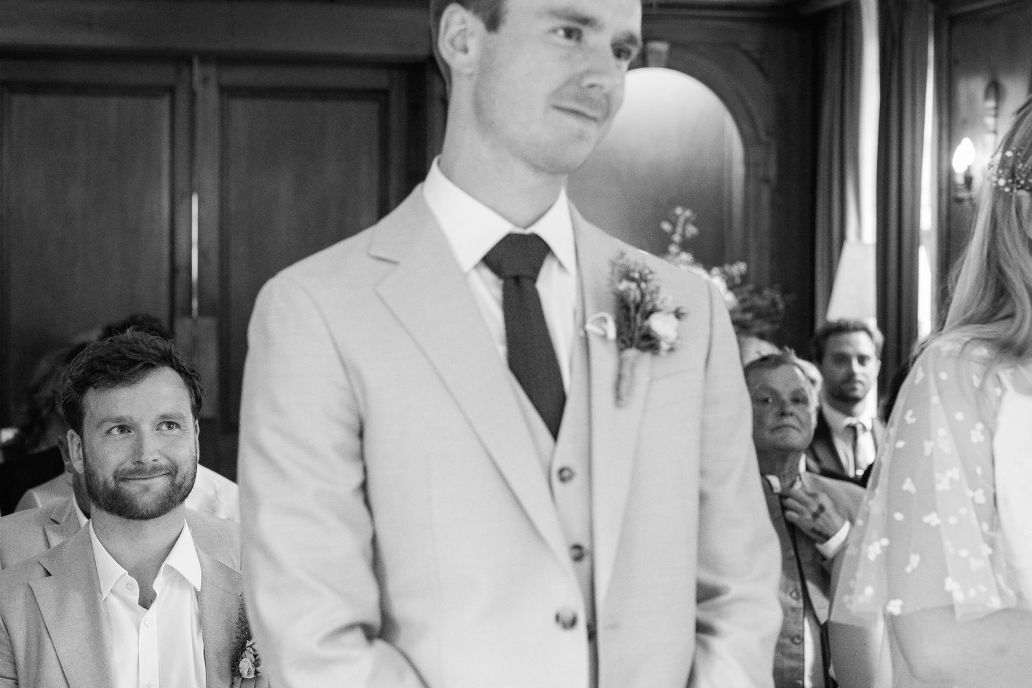 Groom in a light suit with a floral boutonniere stands at the altar while smiling wedding guests look on in a black and white photo.